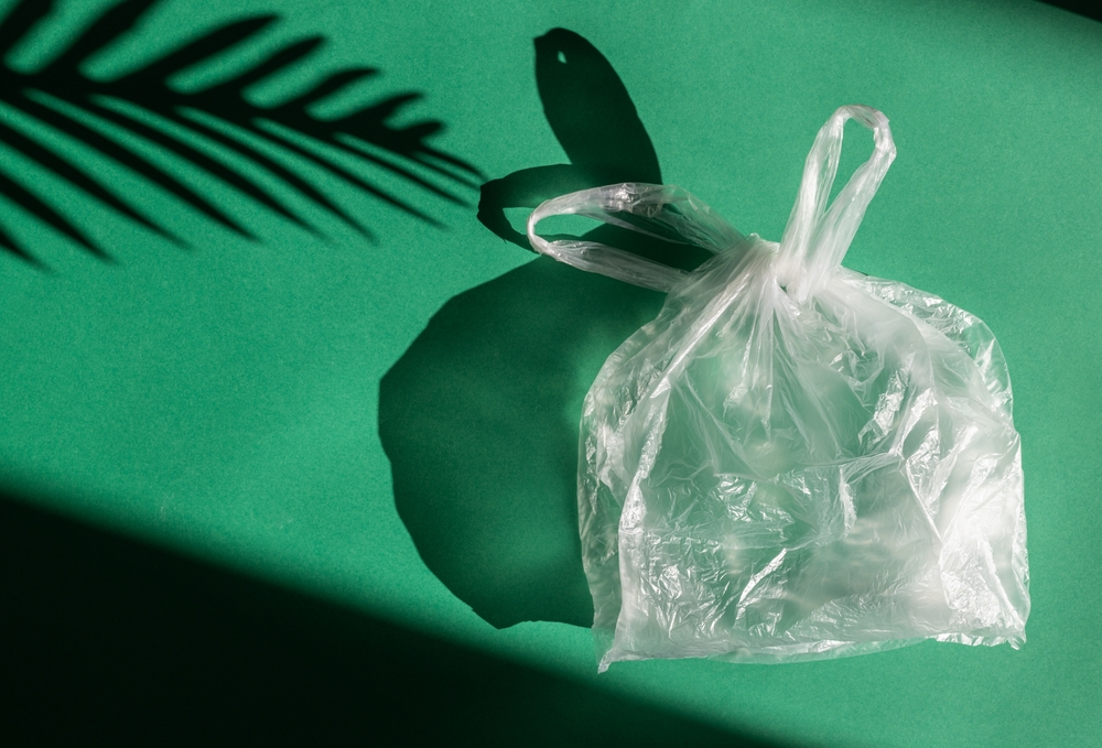 One transparent inflated and tied plastic bag lies on the right on a green background with a hard shadow from the sun, flat lay close-up. Earth day concept, recycling, pollution.