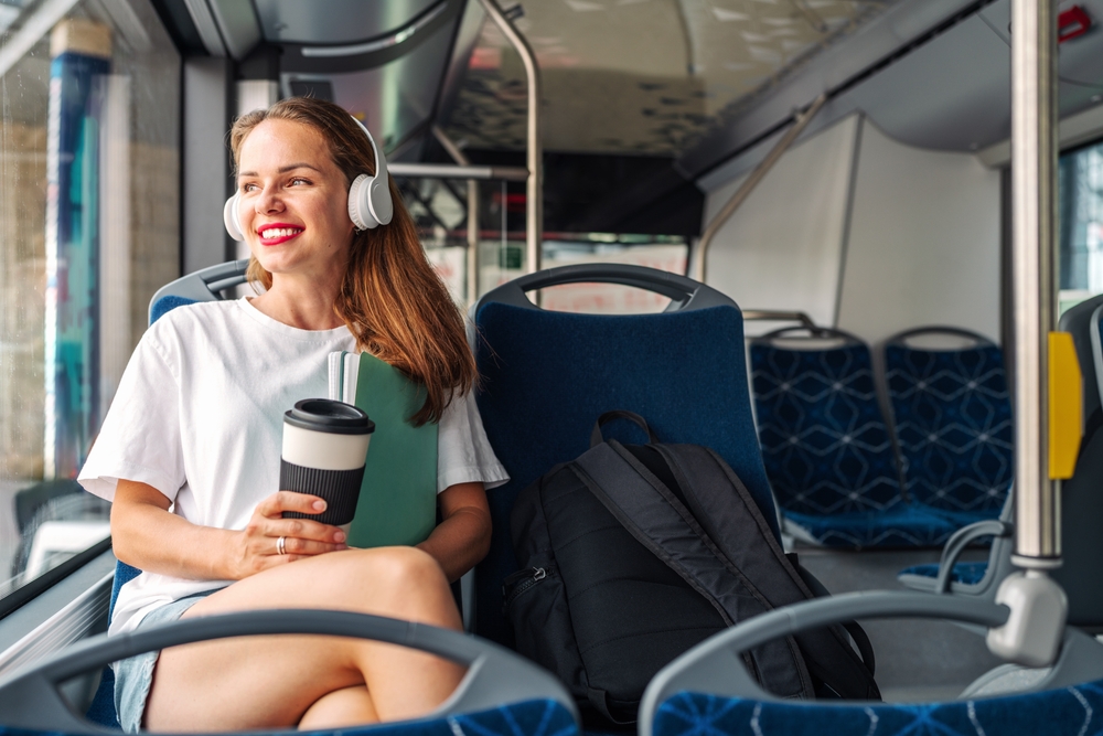 Attractive young adult woman listening music while traveling by city shuttle bus, holding travel mug in hands.