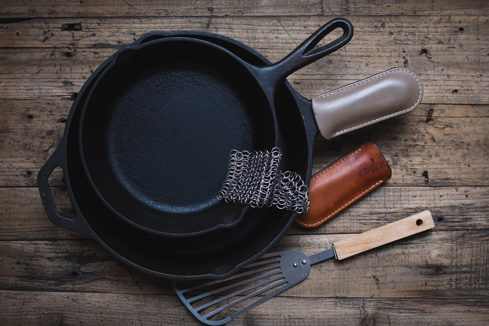 Cast iron skillets, with a metal spatula, leather handle sleeves and a chainmail scrubber on a wooden background. Cast iron cleaning and care.