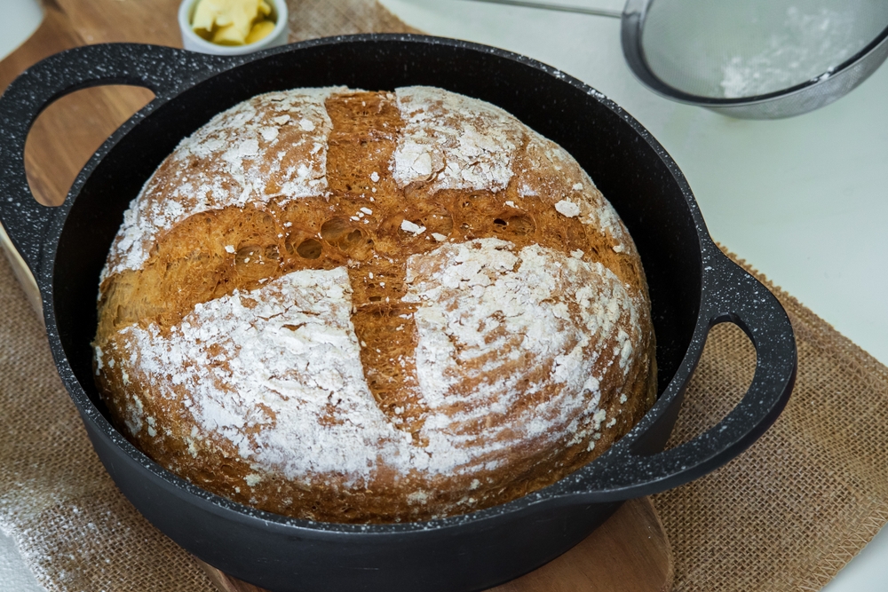 Fresh organic homemade sourdough bread loaf with marks of bread proofing basket inside a cast iron skillet. Golden artisan bread crust with butter on the side on a wooden board. Healthy eating concept