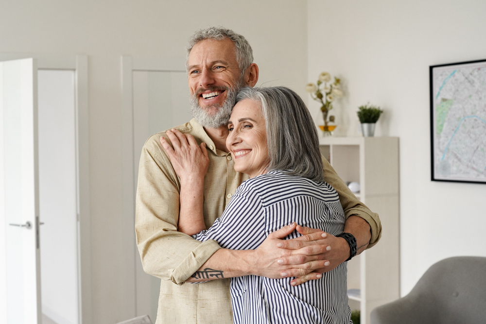 Happy senior adult mature classy couple hugging, bonding, thinking of good future. Carefree cheerful mid age old husband embracing wife looking away dreaming, enjoying wellbeing and love in new house.