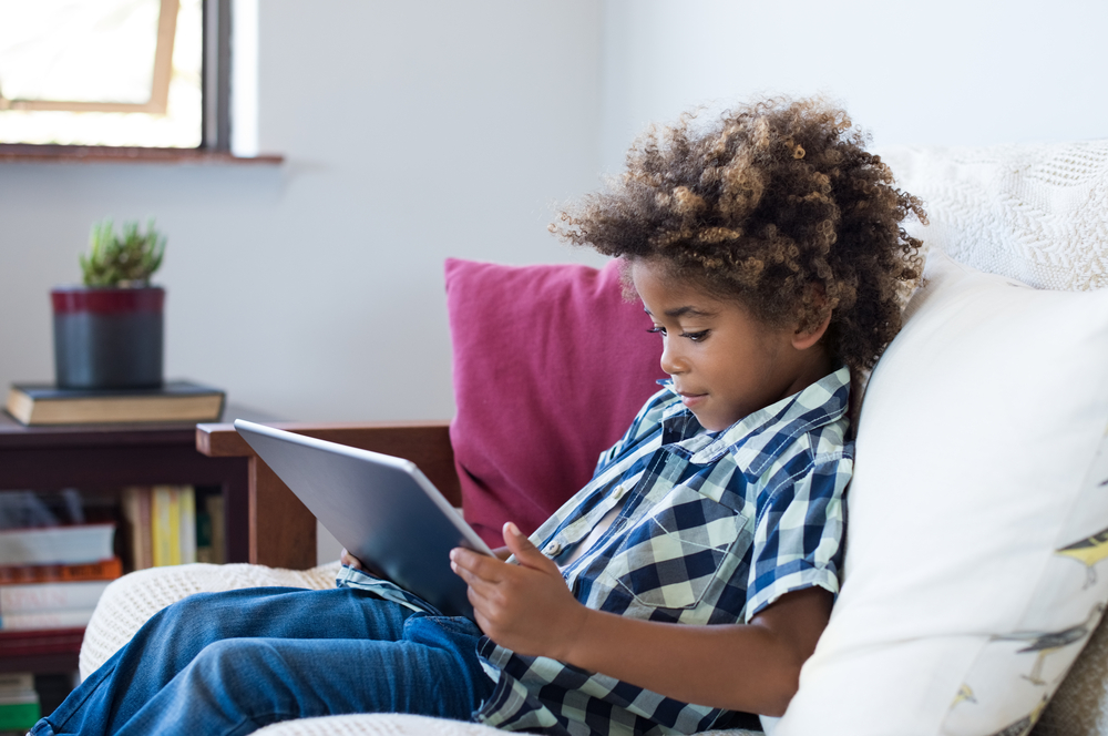 Little african boy sitting on sofa and playing game on digital tablet. Portrait of a young black child at home watching cartoon on the laptop. Modern kid and education technology.