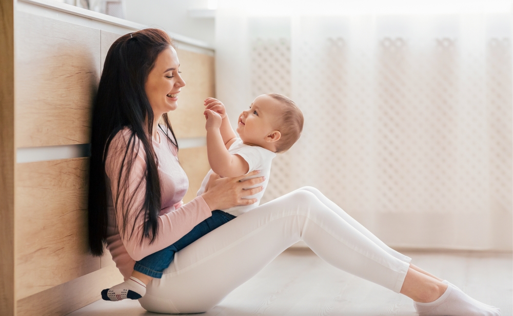 A woman seated on the floor, cradling a baby in her arms. The woman face is attentive, while the baby looks content and safe in her embrace.