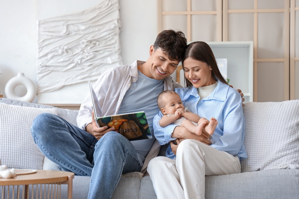 Young parents with their little baby reading book on sofa at home