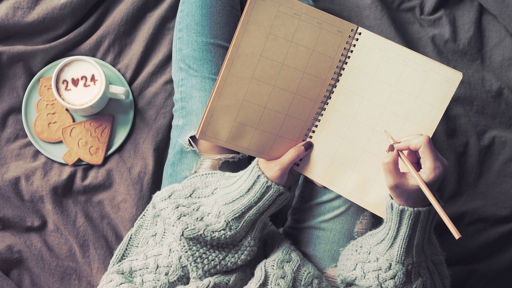 New Year's goal setting, woman in green knitted sweater with jeans write down her resolutions. Notebook and pencil in female hands holding over blurred coffee cup with number 2024 on frothy surface.