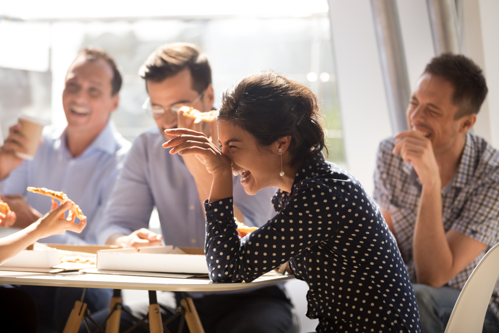 Indian woman laughing at funny joke eating pizza with diverse coworkers in office, friendly work team enjoying positive emotions and lunch together, happy colleagues staff group having fun at break
