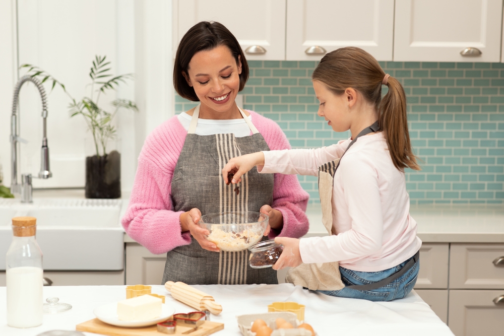 Glad caucasian young mother and little daughter make cookie dough, enjoy prepare eat at spare time in kitchen interior. Homemade food, hobby, cooking sweets together at home
