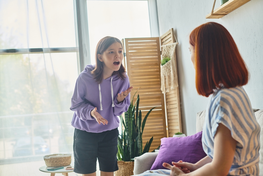 irritated teenage girl screaming and quarreling with mother sitting in living room, family trouble