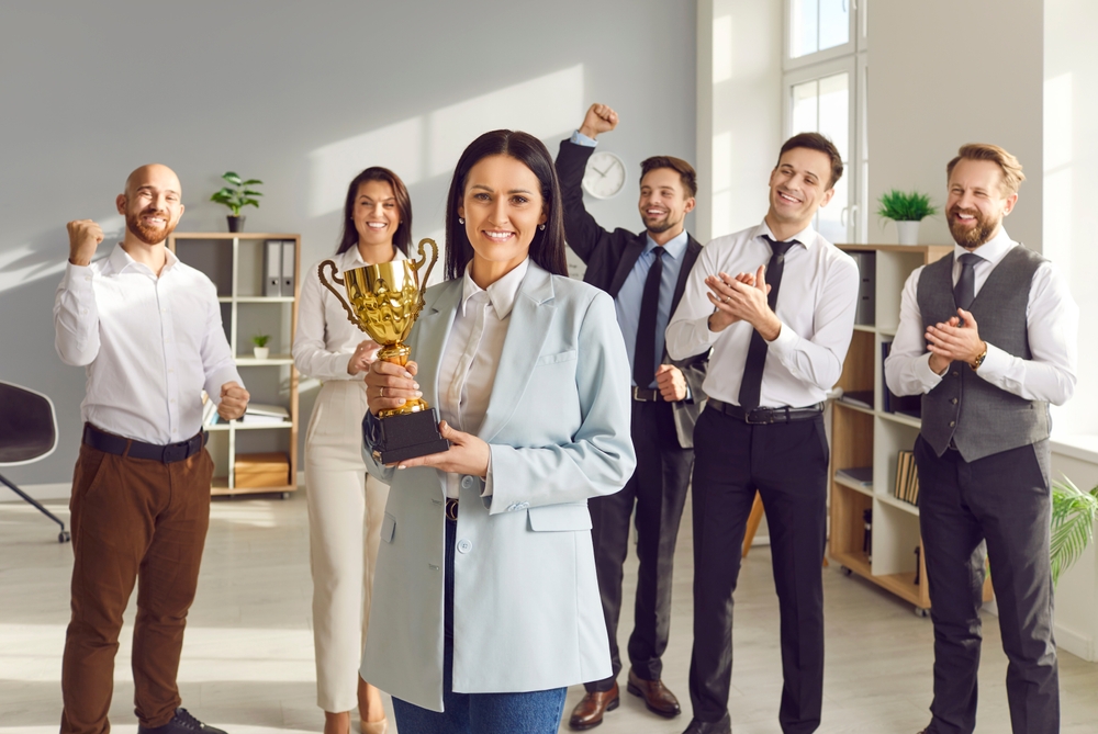 Portrait of a happy smiling young woman winner holding the trophy award for success in business standing in office with a team of people colleagues applauding for company best employee reward.
