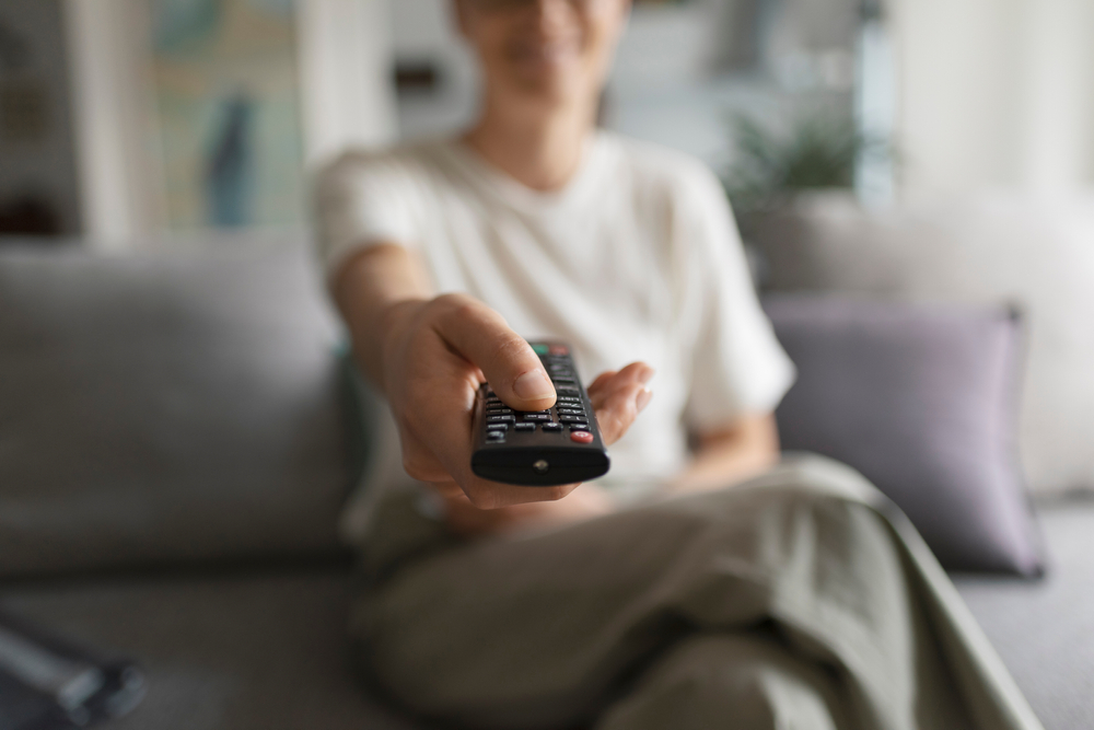 Happy woman sitting on the couch and watching television, she is pointing the remote control at the camera
