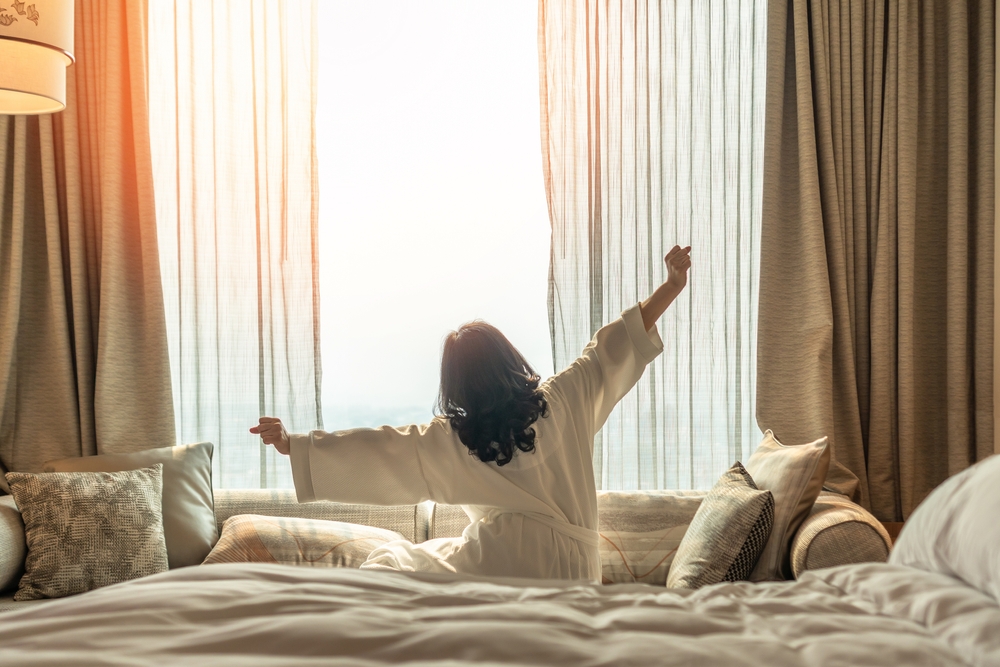 Hotel relaxation on lazy day with Asian woman waking up from good sleep on bed in weekend morning resting in comfort bedroom looking toward city view, having happy, work-life quality balance lifestyle
