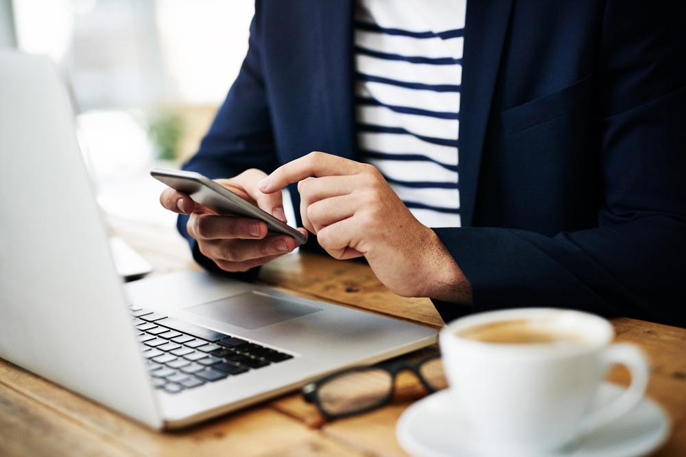 Laptop, hands and businessman with cellphone in office for online email communication for corporate review. Technology coffee and hr with computer and phone for reading company policy on internet,
