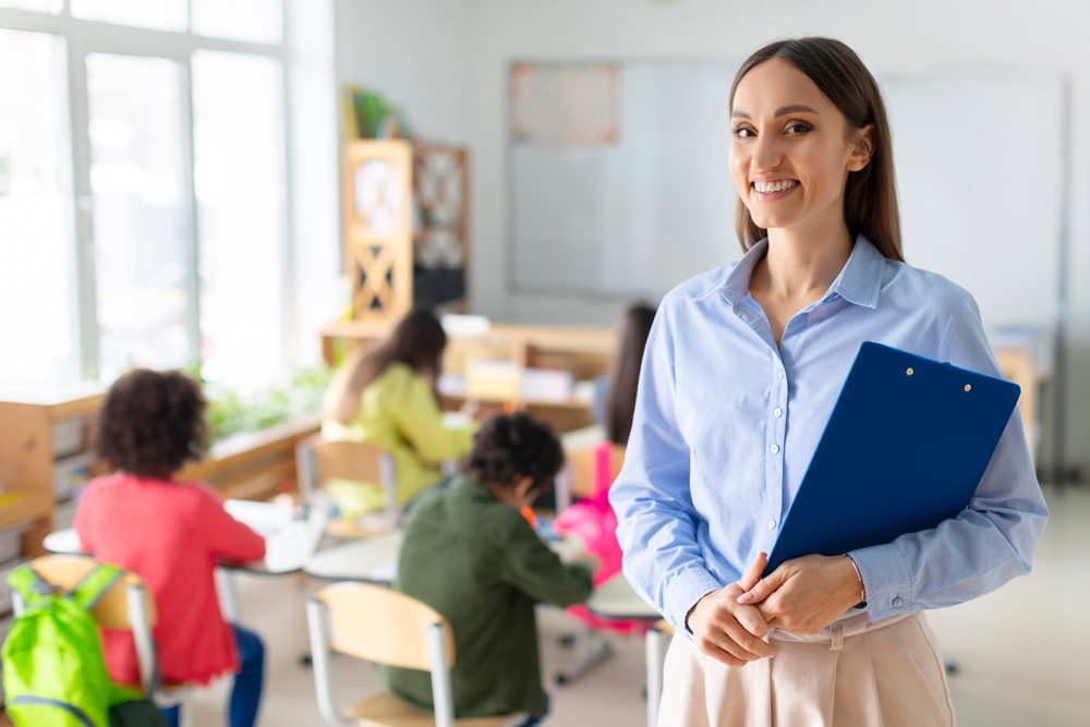Portrait of cheerful European female teacher standing in classroom during lesson with pupils studying on background