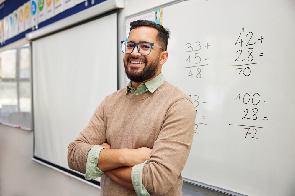 Happy teacher explaining mathematics at whiteboard in classroom. Portrait of smiling mathematics tutor explaining addition concept to children in classroom. Successful teacher standing in classroom.