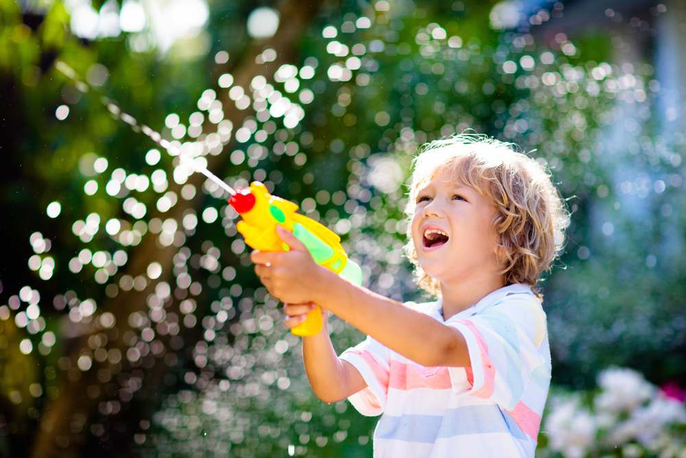 Kids play with water gun toy in garden. Outdoor summer fun. Little boy playing with water hose in sunny backyard. Party game for children. Healthy activity for hot sunny day.
