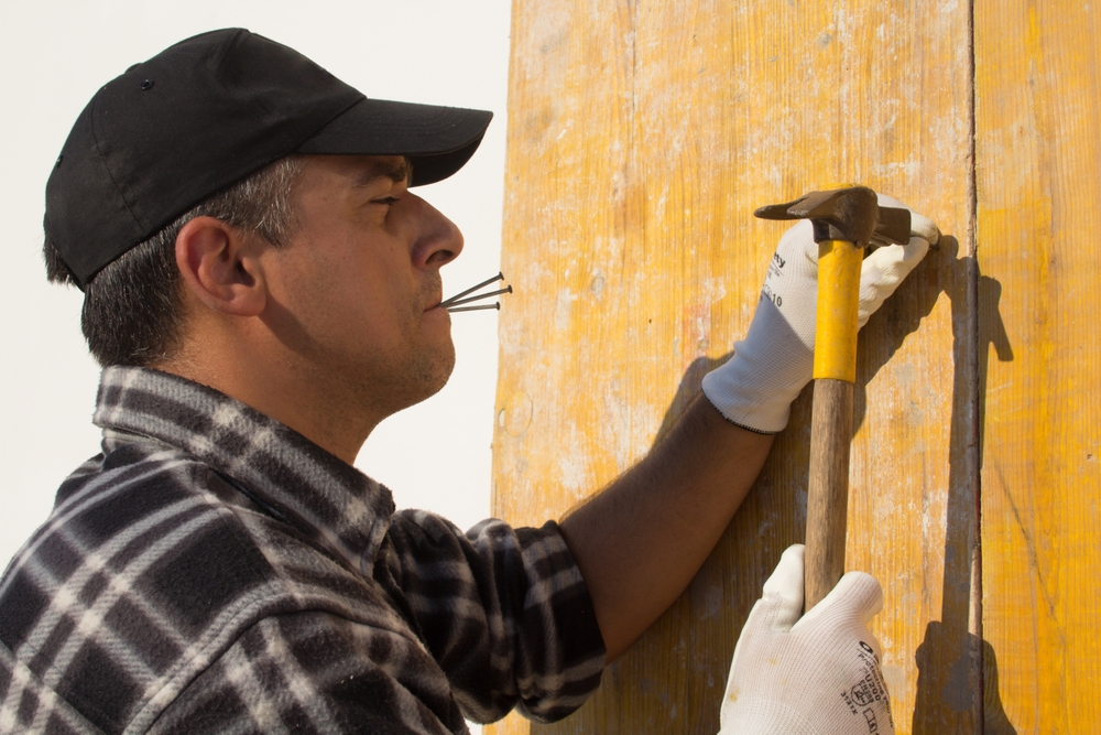 Image of a construction worker with hat and nails between his lips who with a hammer fixes wooden boards. Masonry and construction works
