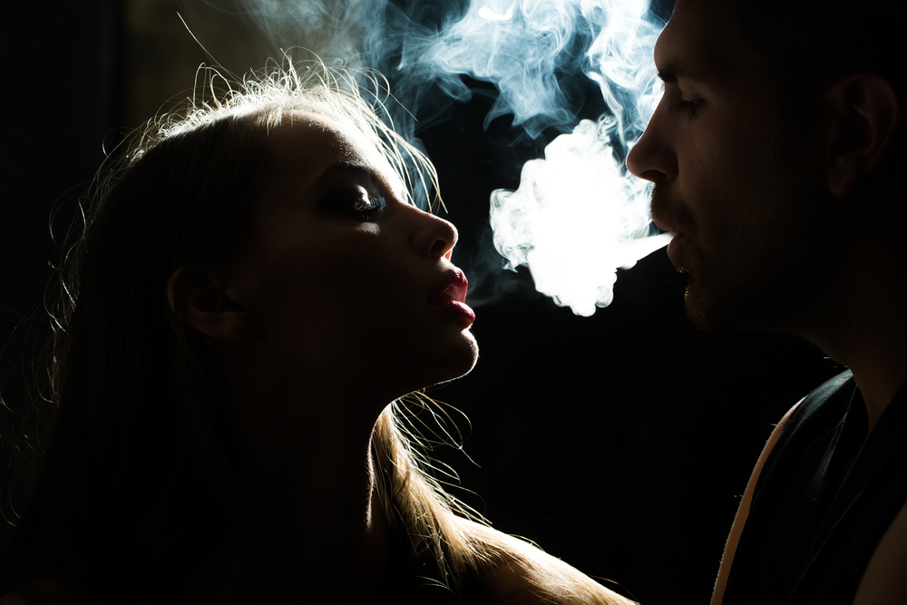 Sensual couple puffing smoke into face beautiful young woman with eyes closed and bearded man looking at her lips in studio play of light and shadow on dark background, horizontal picture