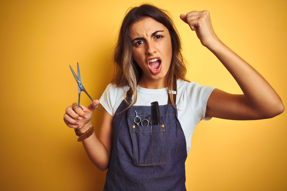 Young beautiful hairdresser woman holding scissors over yellow isolated background annoyed and frustrated shouting with anger, crazy and yelling with raised hand, anger concept