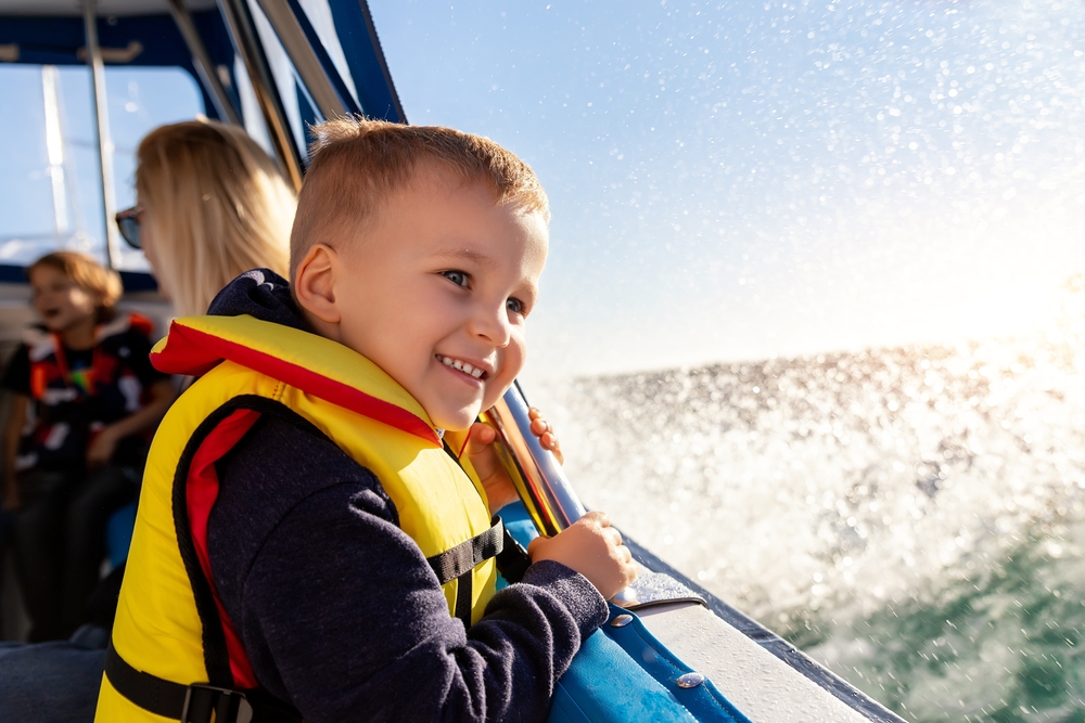 Portrait of cite little blond happy excited smiling caucasian boy wear lifevest enjoy sailing on motor boat sea against blue sky and water splash wave sun backlit. Summer travel vacation recreation