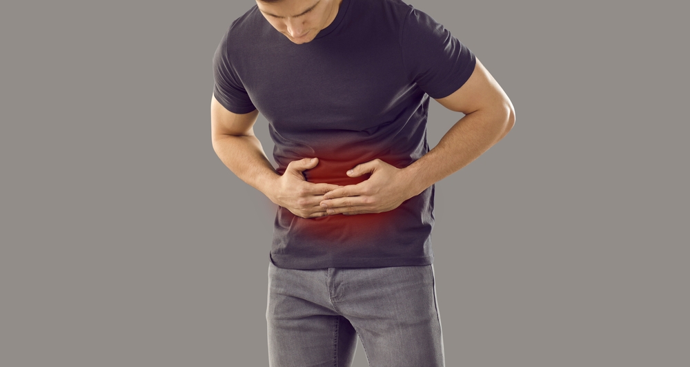 Adult man suffering from digestive disorder. Sick young guy standing isolated on grey banner background feels gastrointestinal discomfort and abdominal pain and touching highlighted red stomach area
