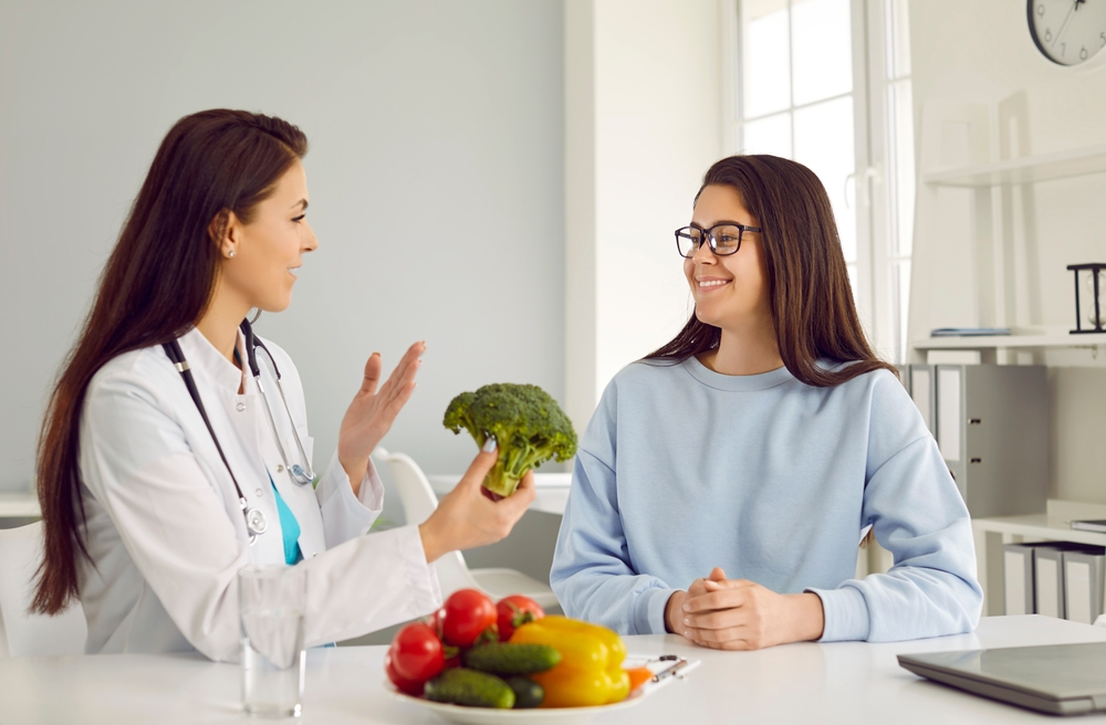 Female nutritionist tells young woman about benefits of eating vegetables during consultation. Smiling woman listens to qualified healthy food expert who holds broccoli in her hands and speaks.