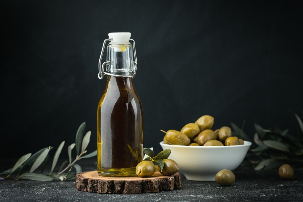 Green olives in a white bowl next to a bottle with olive oil and leaves on a black background. Bottle of cold pressed oil. Traditional Greek and Italian food.
