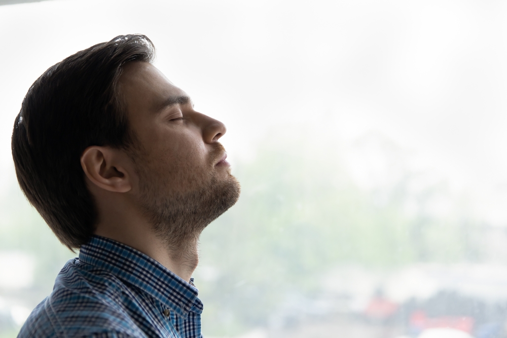 Relaxed millennial 30s man enjoying break at home, taking deep breath of fresh air with closed eyes at window, doing breathing exercises, meditation, relaxing mind, feeling stress relief. Side view