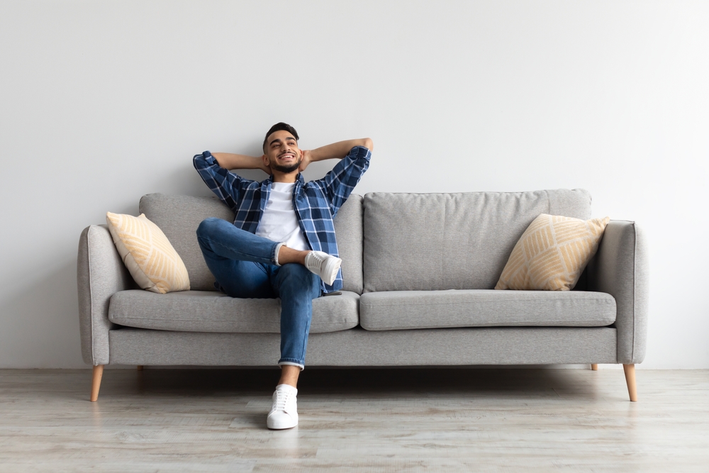 Rest Concept. Happy Middle Eastern guy sitting on comfortable couch at home in living room. Cheerful casual man relaxing on sofa, leaning back, enjoying weekend free time or break from work