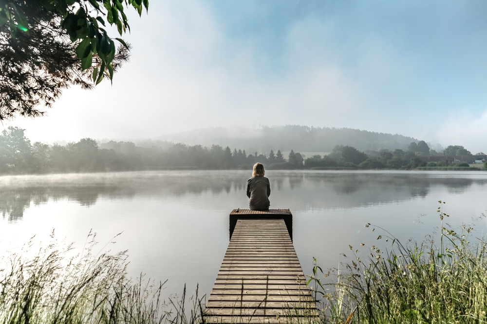 Calm misty morning meditation by pond.Sitting woman outdoors.Wellbeing and wellness soul concept.Spring foggy nature.Silence.Woman feeling freedom,enjoying vacation.No stress,calm mind,relax