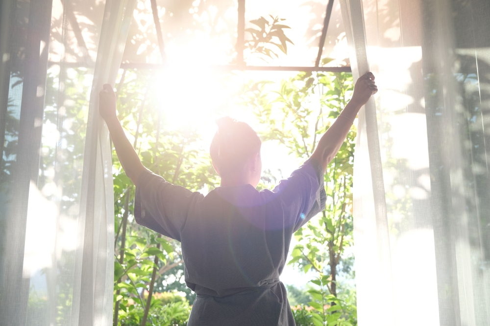 The woman was at the window in the bedroom. She opened the curtains on the window. In the morning and she looks at the view of mountains and trees at sunrise.