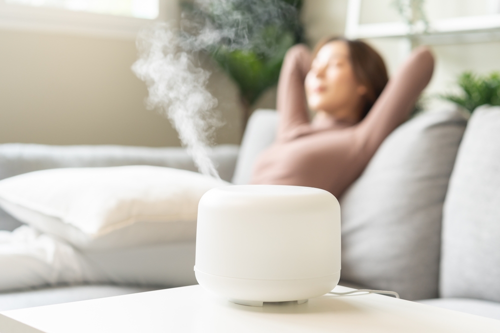 Modern air humidifier during relax or rest, happy blurred asian young woman, girl enjoying aromatherapy steam scent from essential oil diffuser comfortable in living conditions room,apartment at home