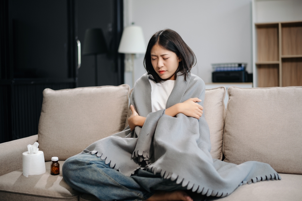 Young Asian woman suffering from flu symptoms, covered with a blanket on a sofa. Concept of illness, healthcare, and recovery. Stay indoors, stay healthy