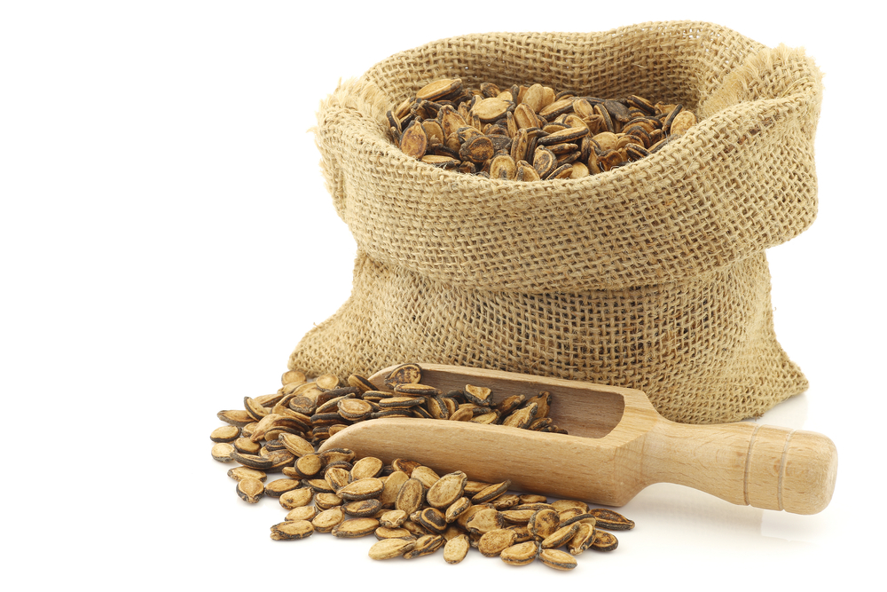 dried watermelon seeds in a burlap bag with an wooden scoop on a white background
