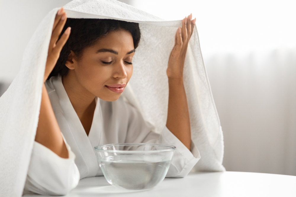 Beauty Hacks. Young black woman inhalling steam of hot water at home, beautiful african american female sitting at table and breathing warm air coming from bowl, covered with bath towel, closeup