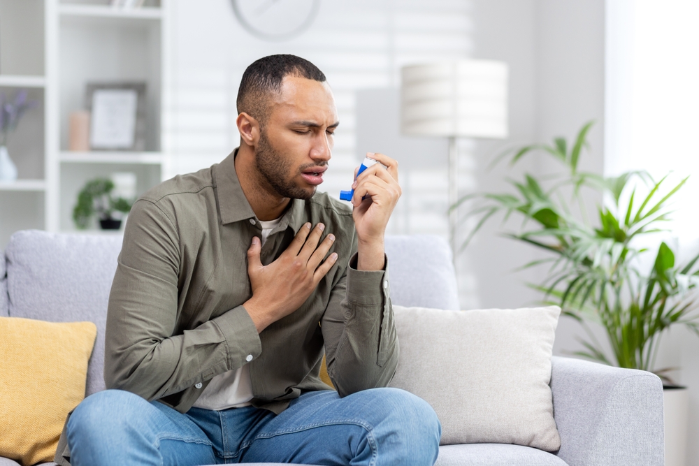 A young African American man is sitting on the couch at home and having an asthma attack. Holds his chest and breathes hard, blows an inhaler into his mouth.