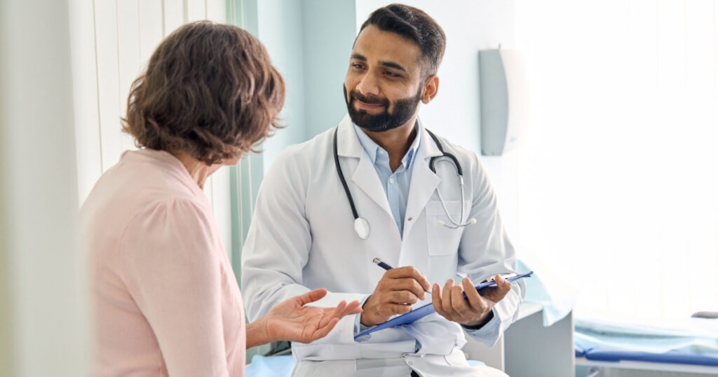 Professional physician wearing white coat talking to mature woman signing medical paper at appointment visit in clinic