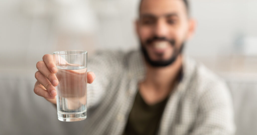 Young Arab man showing glass of fresh clear water at camera at home, selective focus. Millennial middle Eastern guy leading healthy lifestyle, keeping hydrated indoors, copy space. Wellness concept
