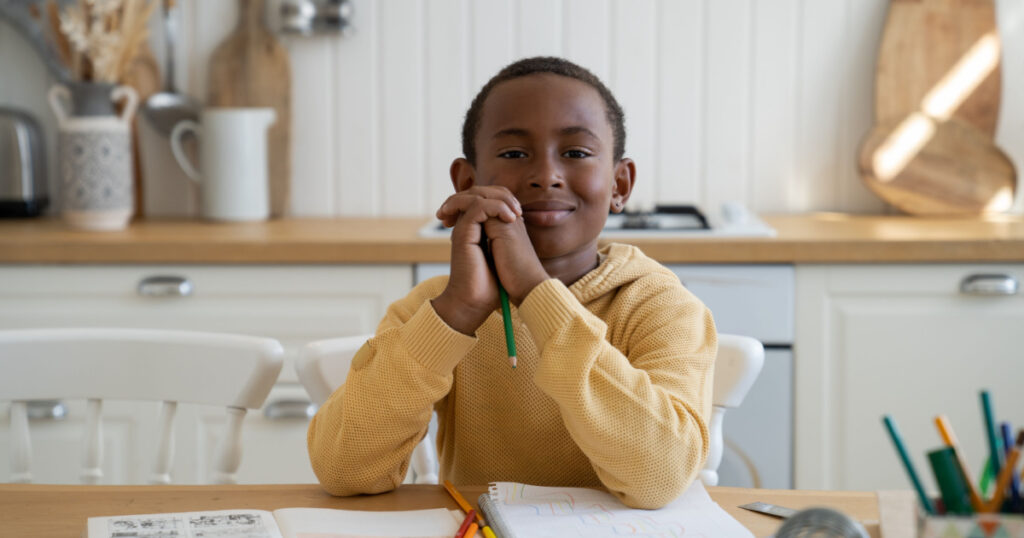 Happy Black schoolboy learning at home with textbook. Smiling African American kid boy sits at desk with pencil look at camera enjoy doing lessons in living room. Son studies while parents at work