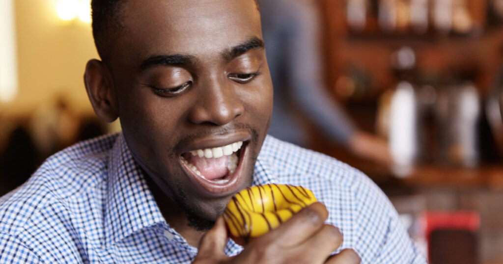 Close-up shot of handsome young dark-skinned man in checkered shirt having tempted look opening his mouth widely, ready to bite doughnut during lunch at restaurant. People, food and leisure concept