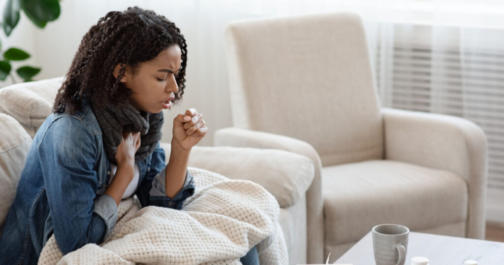 Respiratory illness. Sick black woman coughing hard at home, sitting alone on couch covered with blanket, side view with copy space
