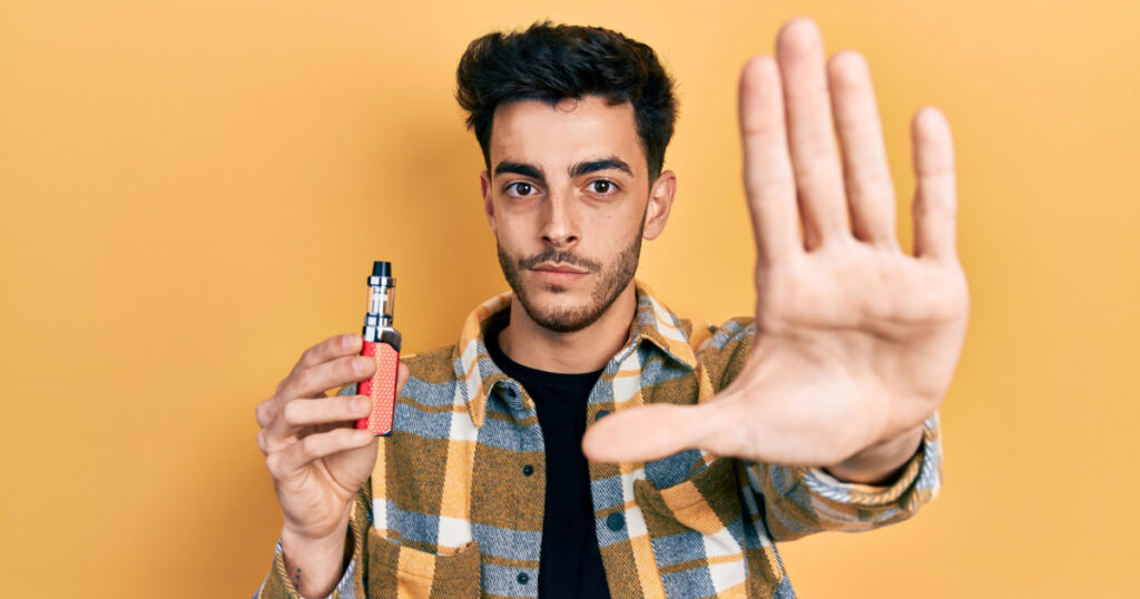Young hispanic man holding electronic cigarette with open hand doing stop sign with serious and confident expression, defense gesture