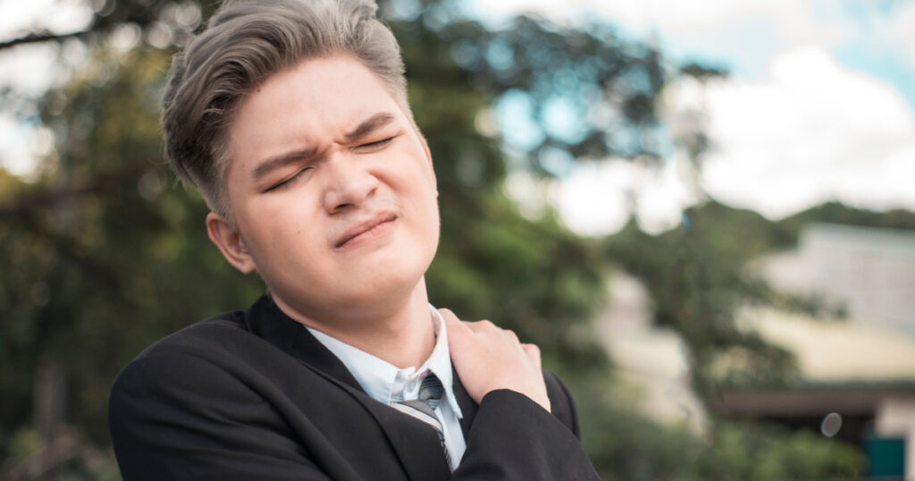 A young man experiences excruciating shoulder pain or stiff trapezius while taking a walk outdoors.