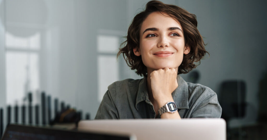 Image of young beautiful joyful woman smiling while working with laptop in office