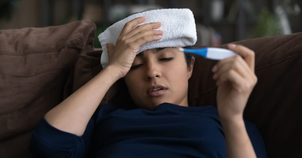 Unhealthy millennial Indian woman struggle with high temperature measure with thermometer. Sick ill young ethnic female suffer from flu or cold, have covid-19 lying at home. Corona virus concept.
