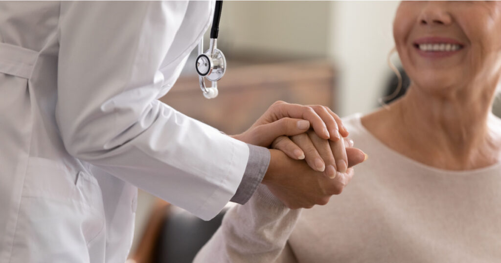 Doctor giving hope. Close up shot of young female physician leaning forward to smiling elderly lady patient holding her hand in palms. Woman caretaker in white coat supporting encouraging old person
