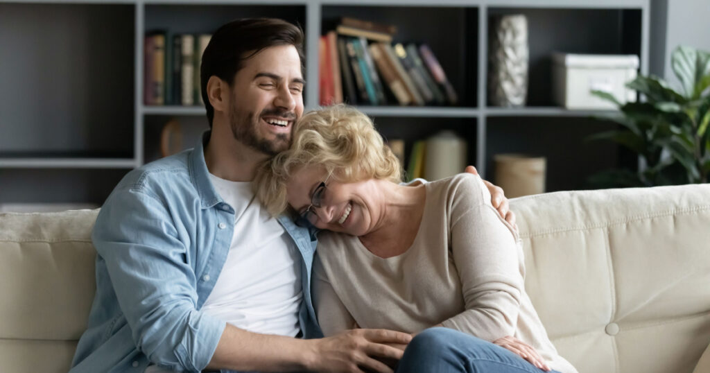 Overjoyed mature senior retired woman leaning on shoulder of happy grown up son, laughing at funny story together while relaxing on comfortable couch in modern living room, weekend family pastime.