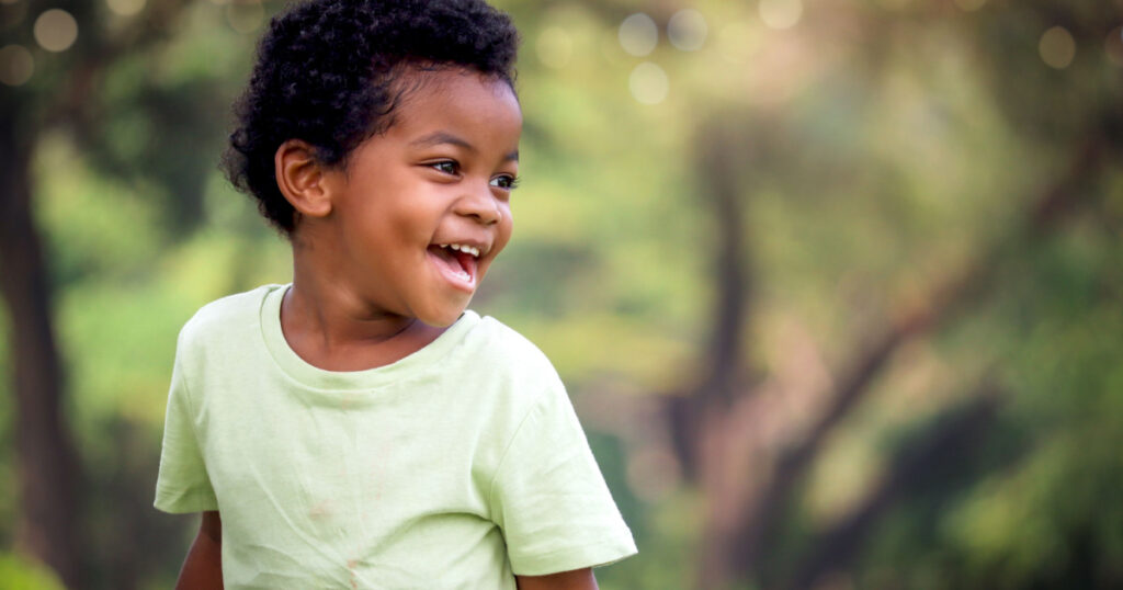 Portrait of happy smiling African boy with black curly hair standing and laughing outdoor with blurred green garden background,