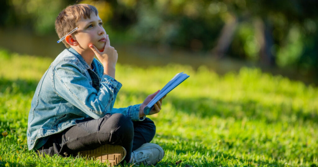 Thinking student with a textbook in his hands is sitting on a green lawn in a park. Young boy reading his notes and memorizing the text before a test. A student writing in a notebook in a garden