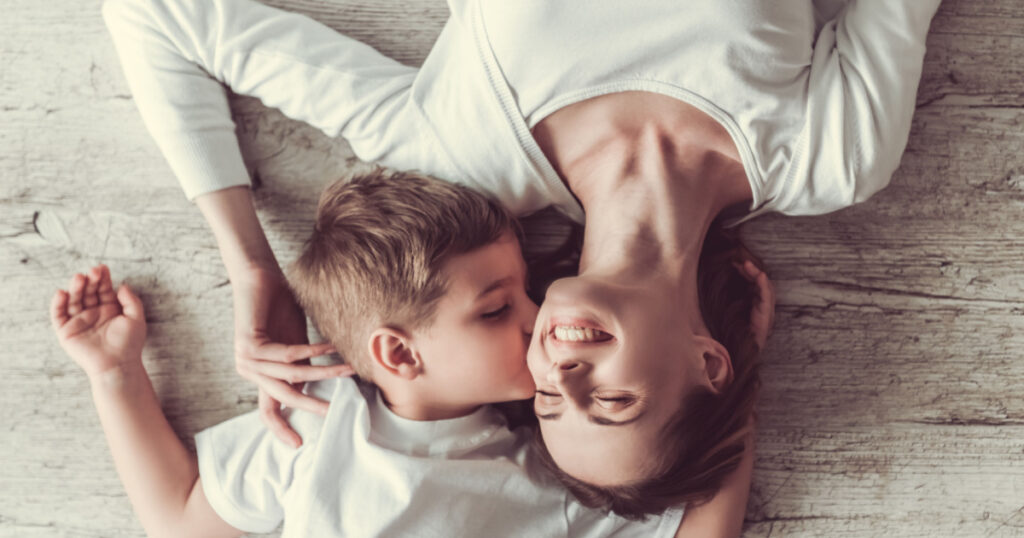Top view of beautiful woman and her cute little son smiling while lying on the floor. Boy is kissing his mom in cheek