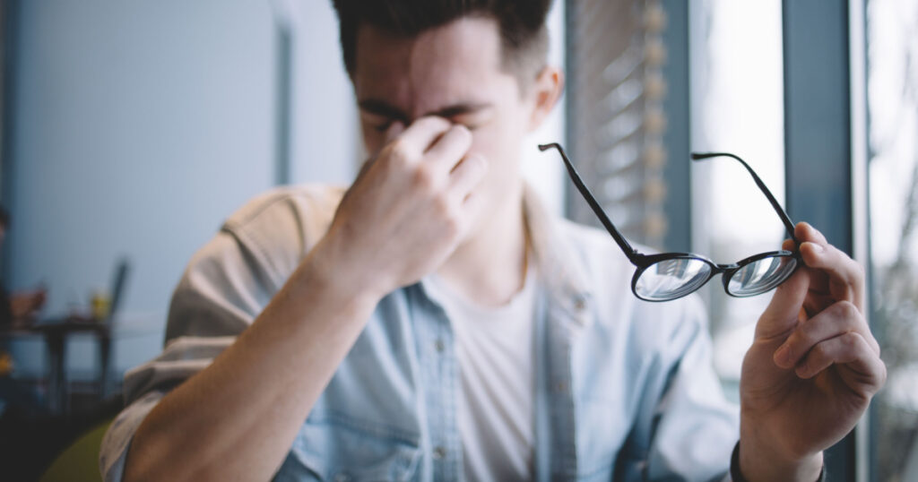 Close up portrait of an attractive man with eyeglasses. Poor young guy has eyesight problems. He is rubbing his nose and eyes because of weariness